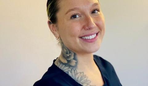 A female with their hair tied back with a tattoo on their neck smiling brightly into the camera