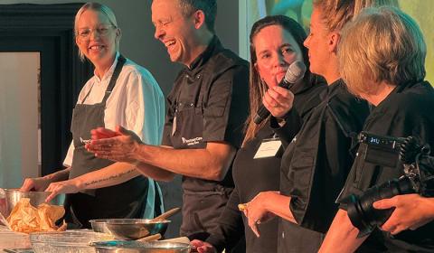 A group of five chefs stands behind a kitchen counter, engaged in a lively cooking demonstration. They are wearing black and white chef uniforms and smiling as they interact with each other. The background features a large screen displaying a food-related image. The atmosphere appears energetic and collaborative, with various cooking utensils and ingredients visible on the counter.