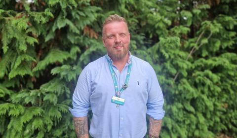 A man with a beard stands in front of a dense green hedge. He is wearing a light blue button-up shirt and has tattoos visible on his arms. A lanyard with a badge hangs around his neck. The background features lush foliage, creating a natural setting.