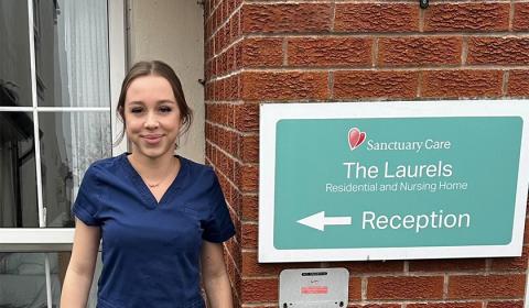 A person in navy blue scrubs standing outside a brick residential and nursing home beside a sign reading ‘Sanctuary Care – The Laurels – Residential and Nursing Home"