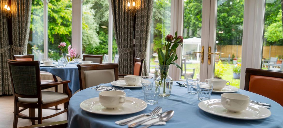 Cosy dining room setup with blue table cloth and chairs, in a room with multiple windows letting in lots of natural light