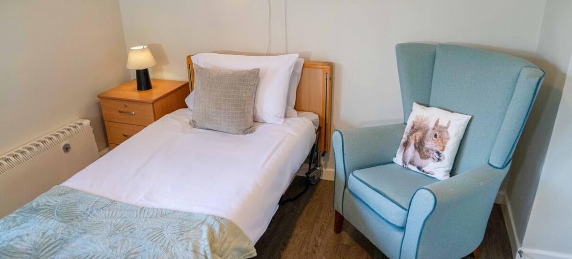 A clean minimal bedroom with a medical single bed with an oversized blue arm chair.