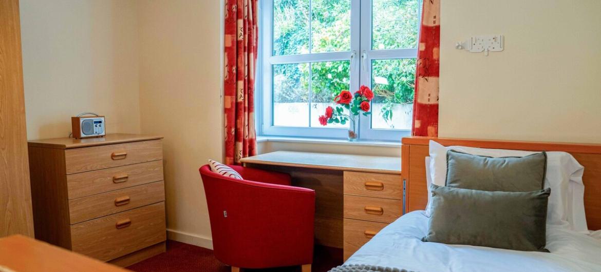 A warm and inviting single bedroom, nostalgic touches like a vintage radio, and a comfy armchair by a sunny window with views of greenery.