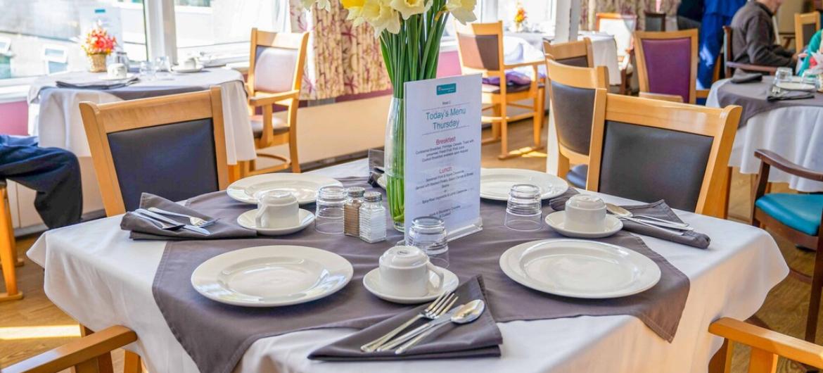 A bright, welcoming dining area at The Woodland, with neatly set tables, fresh flowers, and large windows letting in natural light. Creating a warm, friendly, and community-focused atmosphere.