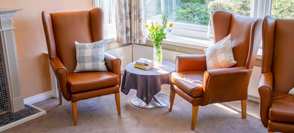 A bright and cosy sitting area, with comfortable large leather armchairs, soft cushions, and a small table topped with fresh flowers and books. Large windows fill the space with natural light, creating a warm and welcoming environment.