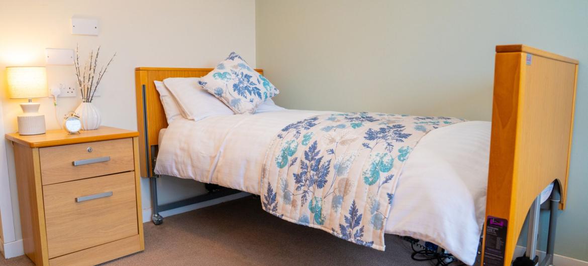 A cosy bedroom scene featuring a neatly made bed with a decorative floral pillow. Beside the bed is a wooden nightstand with a small potted plant and a lamp that emits a warm glow. The walls are painted a soft colour, creating a calm and inviting atmosphere.