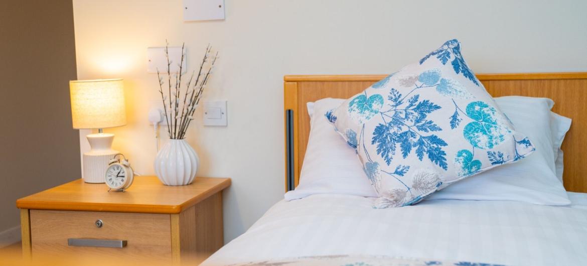 A cosy bedroom scene featuring a neatly made bed with white linens and a decorative pillow with a blue floral pattern. Beside the bed, there is a wooden nightstand with a small clock and a white vase containing dried branches. A soft lamp emits a warm glow, adding to the inviting atmosphere of the room. The walls are painted a light colour, enhancing the overall brightness of the space.