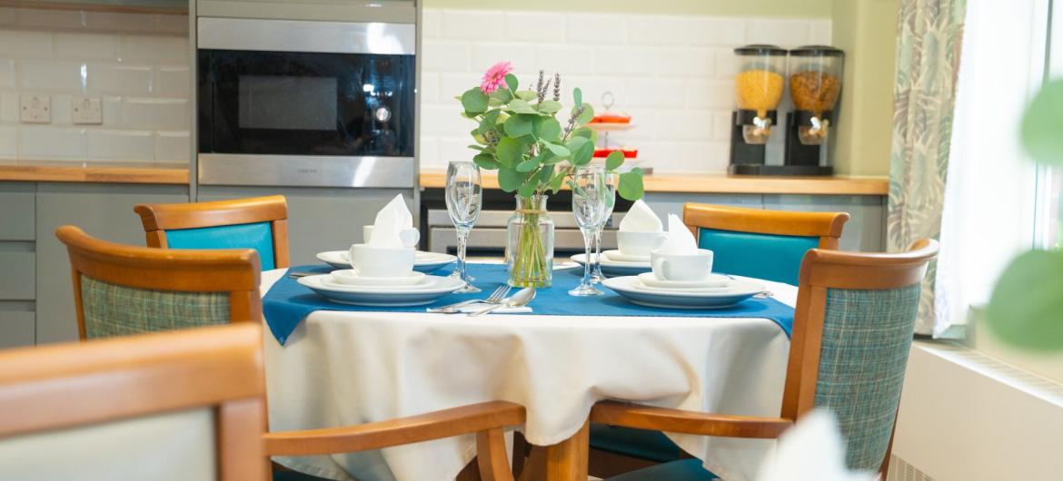 A dining table is elegantly set with white plates, glasses, and folded napkins, all arranged on a blue tablecloth. A small vase with greenery and a pink flower sits at the center. The background features a modern kitchen with a wall clock, an oven, and two glass containers filled with cereal. The chairs are wooden with blue cushions, creating a warm and inviting atmosphere.