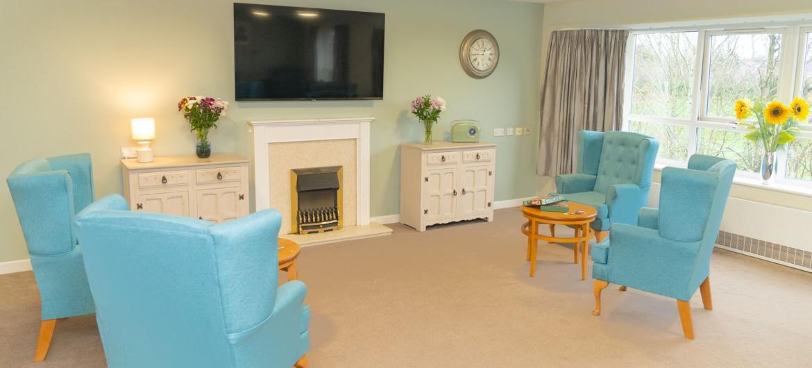 A cosy living room featuring light blue armchairs arranged around a small wooden table. A white fireplace with a decorative mantel is centred on the wall, beneath a large flat-screen TV. To the left, a side table with a lamp and a vase of flowers adds a touch of warmth. A window with sheer curtains allows natural light to fill the space, and a sideboard with decorative items is positioned against the wall. The room has a soft beige carpet and light green walls, creating a welcoming atmosphere.