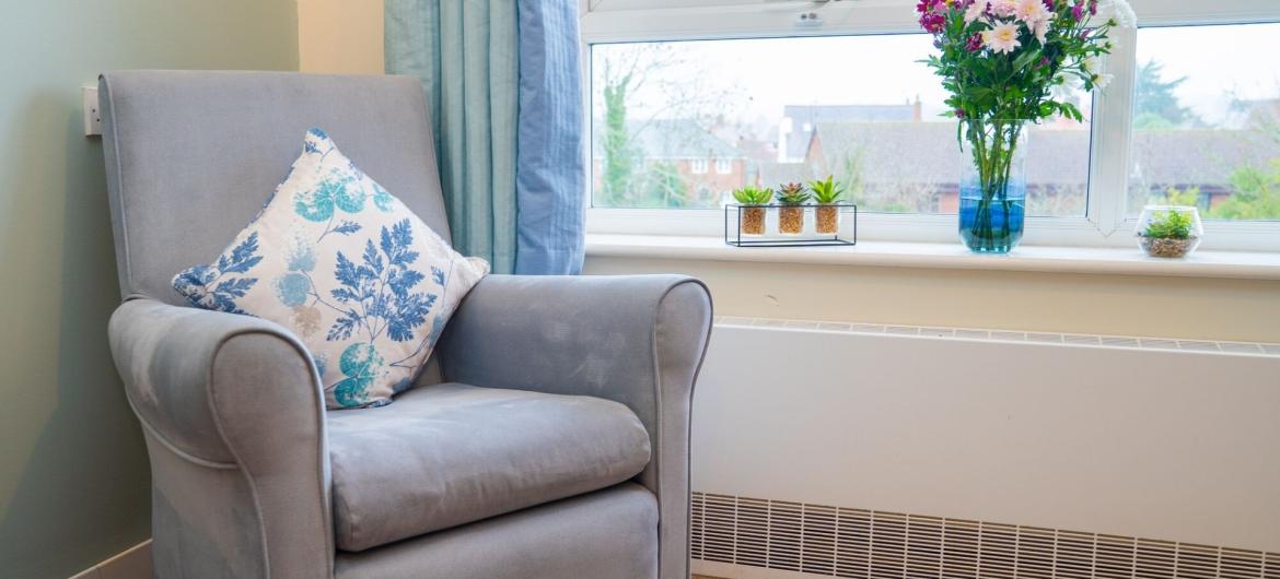 A cosy grey armchair with a decorative blue and white pillow is positioned near a window. The window offers a view of the outdoors, with soft natural light illuminating the space. On the windowsill, there is a vase of colourful flowers and small potted succulents, adding a touch of greenery to the room. The walls are painted a soft green, and the floor is covered with beige carpet.