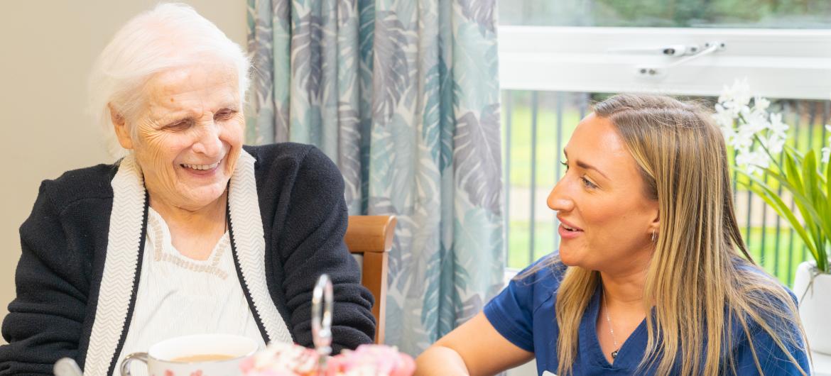 An elderly woman with white hair and a warm smile sits at a table, laughing with a younger woman in a blue uniform. The table is set with a plate of pink frosted cupcakes and cups of tea. In the background, there are large windows with natural light and greenery visible outside, creating a cheerful atmosphere.
