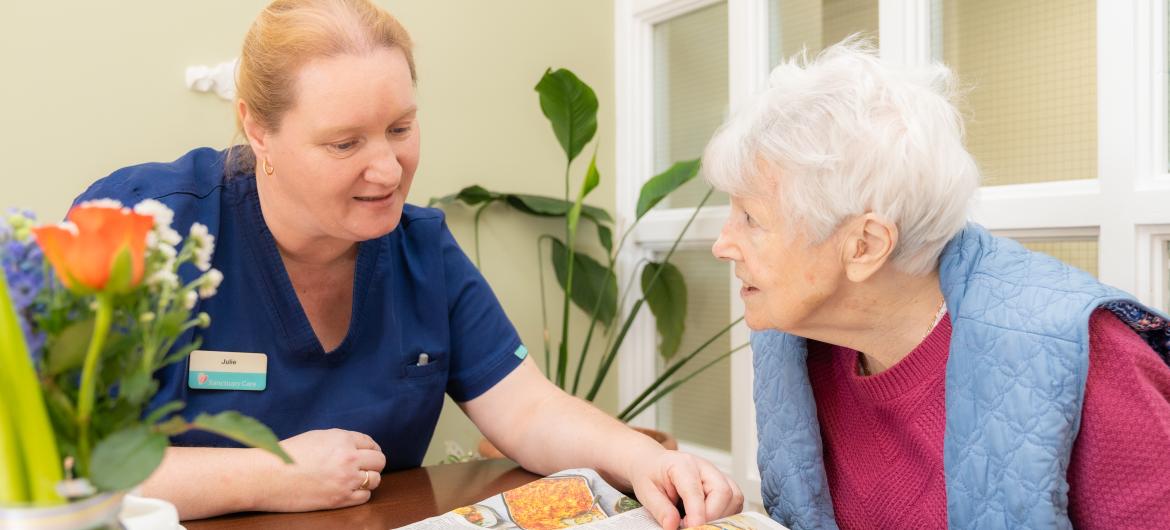 A healthcare worker in a blue uniform is sitting at a table with an elderly woman who has white hair. The worker is pointing at a magazine, engaging the woman in conversation. A vase with flowers and a cup are on the table, and there is a green plant in the background. The setting appears warm and inviting, suggesting a supportive environment.