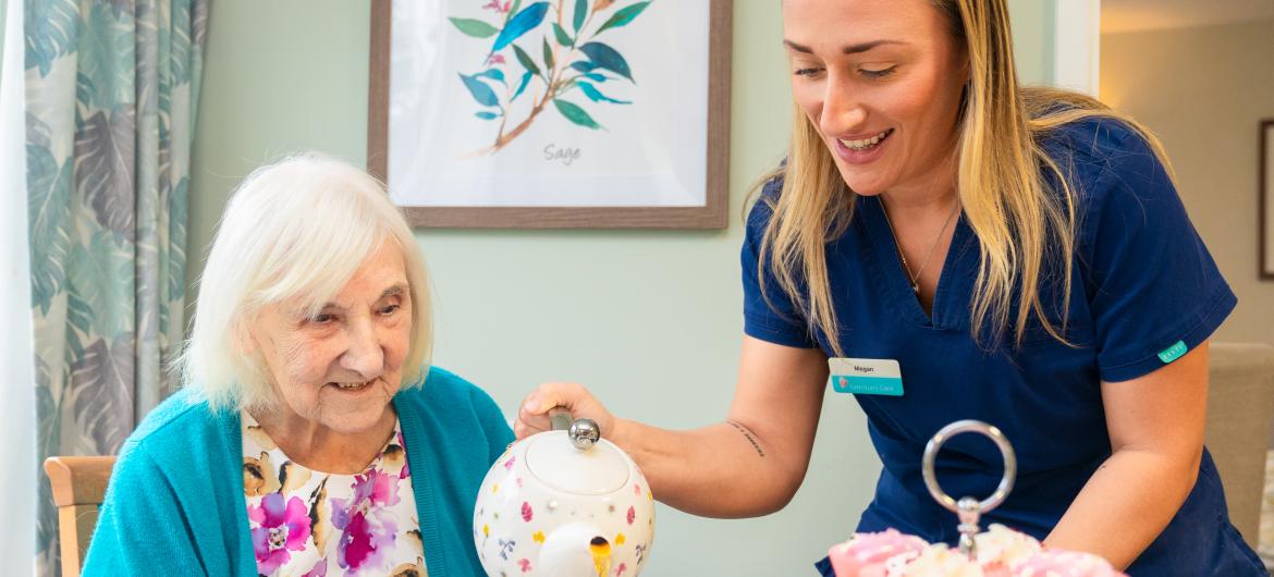 A caregiver is pouring tea from a teapot into a cup for an elderly woman sitting at a table. The elderly woman, with white hair, is smiling and wearing a colorful sweater. The table is set with a floral-patterned tableware, and there are cupcakes displayed on a tiered stand. In the background, there is a framed floral artwork on the wall. The setting appears warm and inviting, suggesting a cozy tea time.