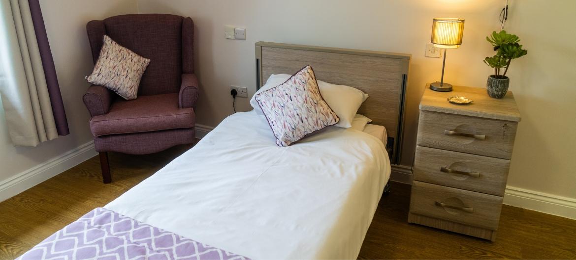 A cosy bedroom with bedside table and lamp, and arm chair