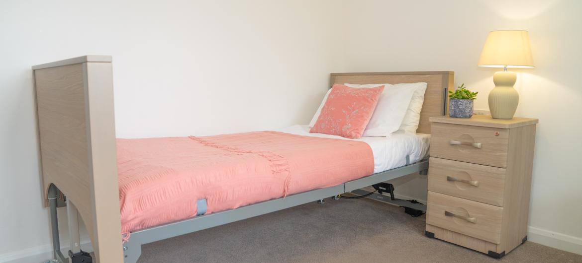 A neatly arranged bedroom featuring a single bed with a light wooden frame and a pink bedspread. The bed is positioned against a white wall. There is a white pillow and a pink decorative pillow on the bed. Next to the bed is a light wooden nightstand with a lamp and a small potted plant on top. The room has a beige carpeted floor, creating a warm and inviting atmosphere.