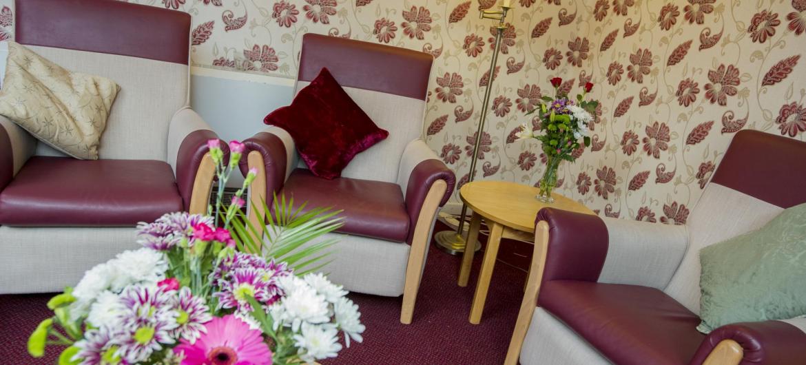 The lounge at Pinewood Residential Care Home with soft chairs, flowers and soft lighting.