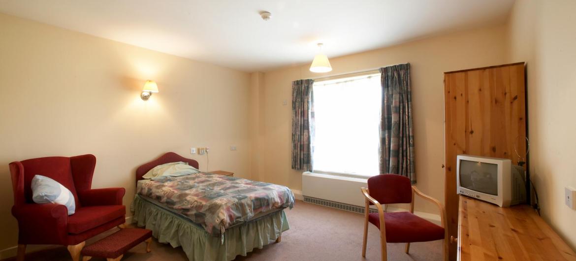 A large bedroom with comfy chair and matching wooden furniture at Meadows House Residential and Nursing Home.