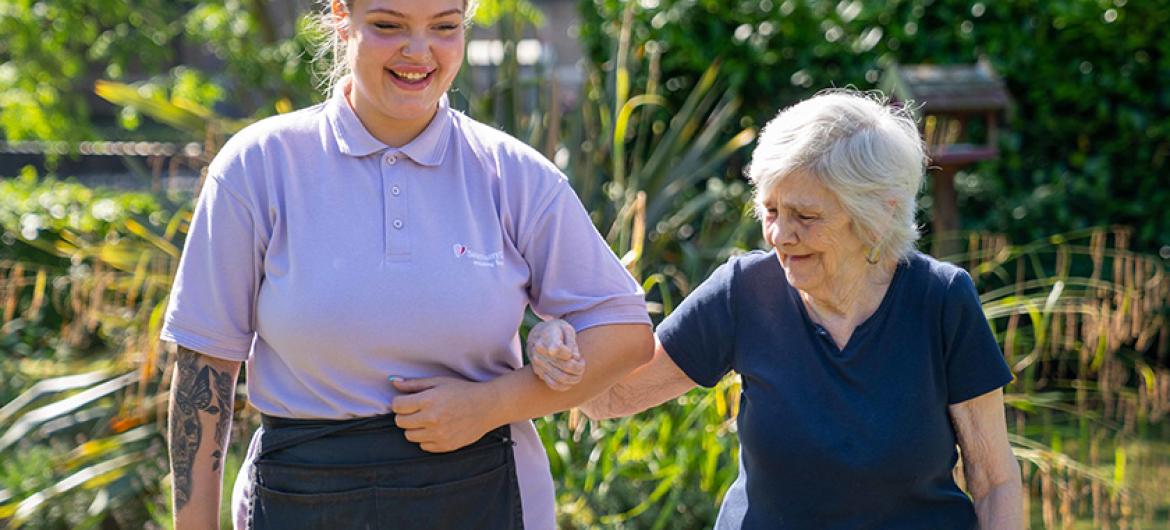 A care worker smiling while guiding an elderly lady through the gardens.