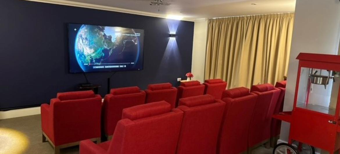 Cinema styled seating room with red plushed arm chairs placed into two rows facing a wall mounted screen, with a dark plum wall paint and black out curtains used. A decorative popcorn machine sits behind the rows of seating.