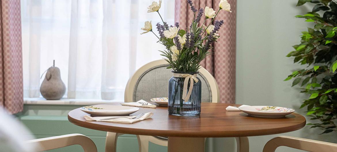 A peaceful seating area with numerous chairs located around the wooden circular dining table. It is decorated with fresh flowers to completement the pale colour pallet of the room.