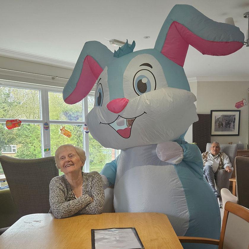 Sanctuary Care resident with the Easter bunny. A female Sanctuary Care resident sits at the table with a giant inflatable Easter bunny, smiling at the surprise.