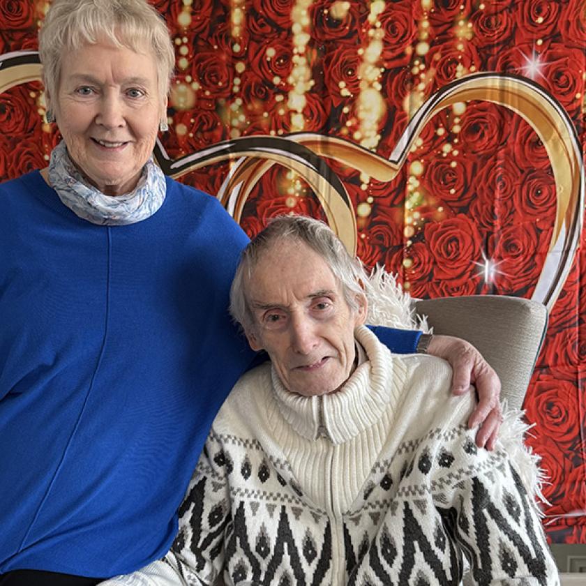 Sanctuary resident Tom sat in an armchair with his wife Clare sat on the arm of the chair in front of a backdrop of roses.