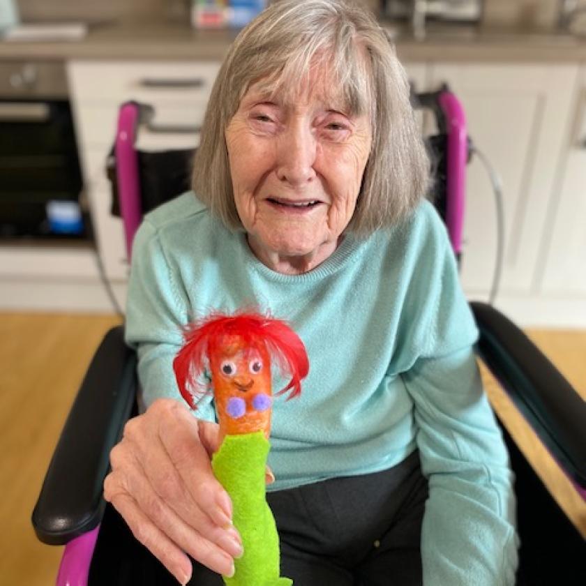 Resident Jo sat in a wheelchair holding a carrot which has been decorated to look like the little mermaid, with green felt tail, red feathered hair, googly eyes and a drawn on smile.
