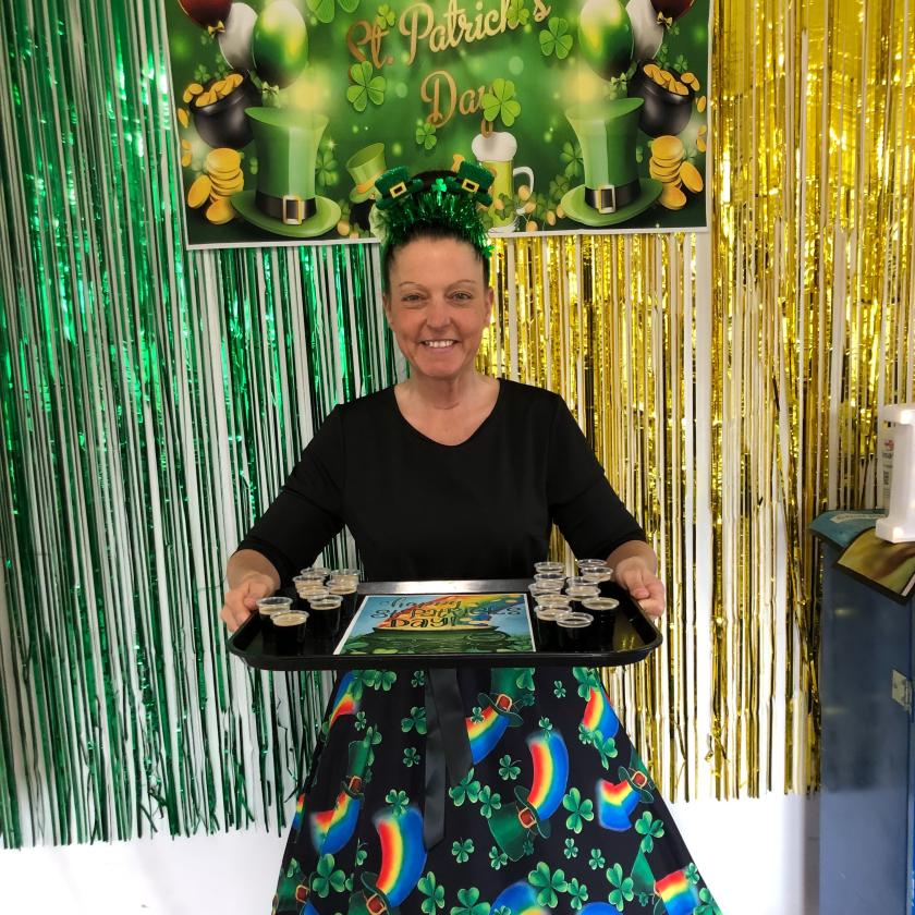 A woman wearing a rainbow skirt holding a tray of Guiness with a Happy St Patricks Day banner behind her