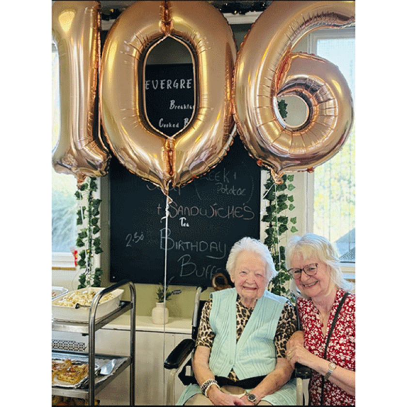 Maria Blagden and Resident Joyce Penfold sitting at The Green Residential Care Home in Redruth Cornwall. They are sat together and smiling with 106th birthday balloons behind them. They are both smiling