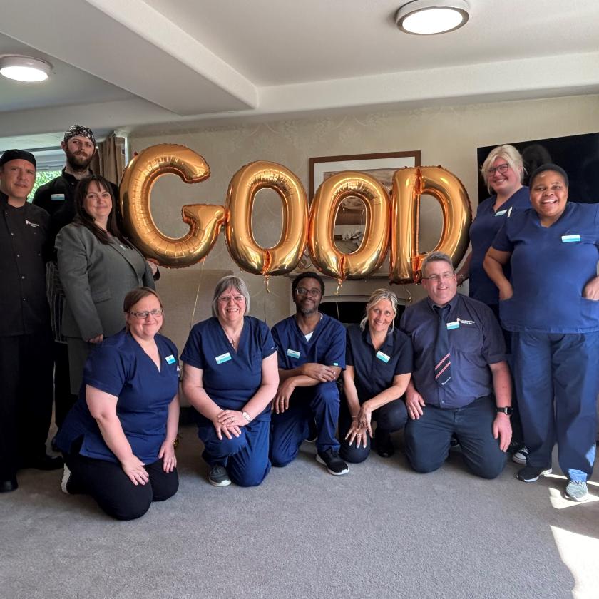 A group of people pose indoors with gold balloons spelling "GOOD"; some are kneeling, others standing, in a bright room with a window and TV.