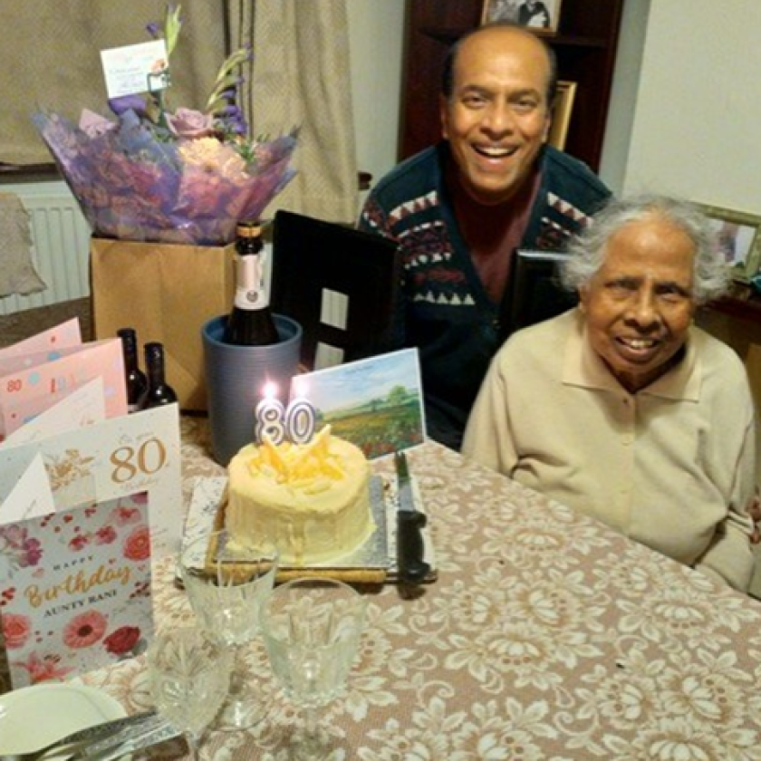 An older person and another individual seated at a table decorated for an 80th birthday celebration, featuring a cake with '80' candles, birthday cards, flowers, and a bottle of champagne.
