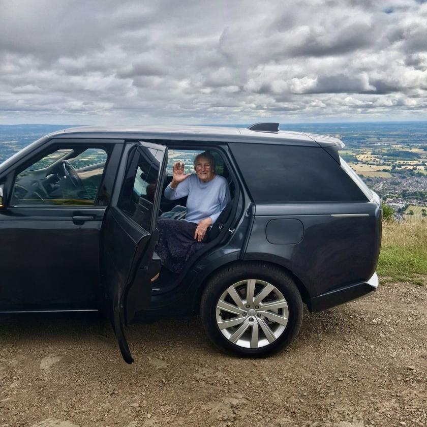 Person seated in the back of a black SUV with the rear door open, waving on a dirt road with scenic landscape and cloudy sky in the background