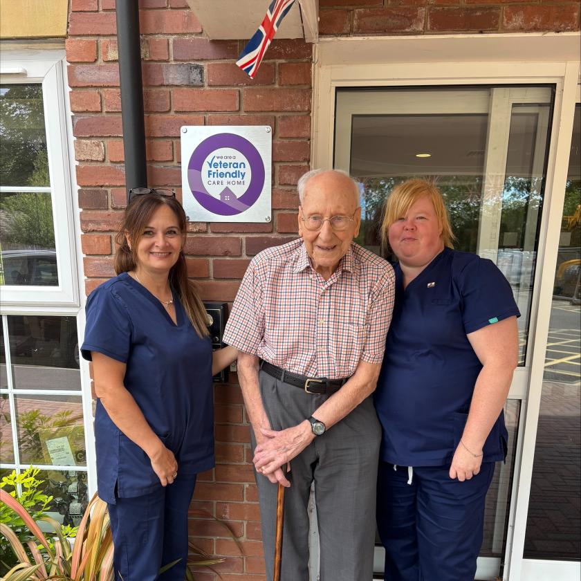 Three people standing outside a building marked 'Veteran Friendly Care Home.' The central person holds a cane, flanked by two in blue uniforms. A British flag is above a purple-circled sign.