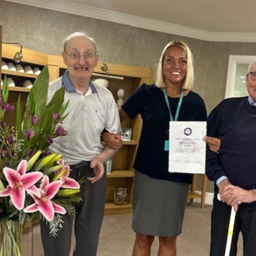 Three people stand indoors, with the person in the middle holding a certificate. A bouquet of flowers sits on a small table to the left. Shelves and a door are visible in the background.