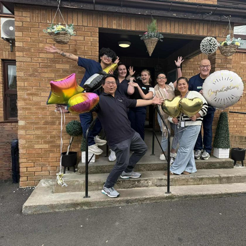 A group of seven people is gathered on the steps of a brick building, celebrating with balloons. They are smiling and posing playfully,