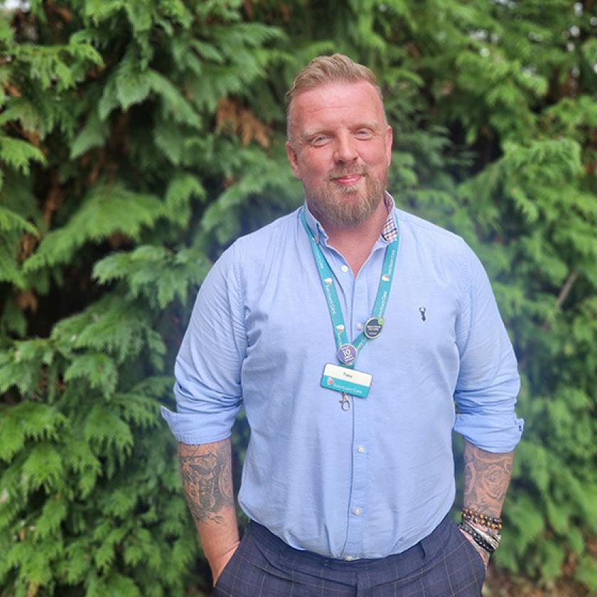 Sanctuary Care Property Manager Tony Kirkby A man with a beard stands in front of a dense green hedge. He is wearing a light blue button-up shirt and has tattoos visible on his arms. A lanyard with a badge hangs around his neck. The background features lush foliage, creating a natural setting.