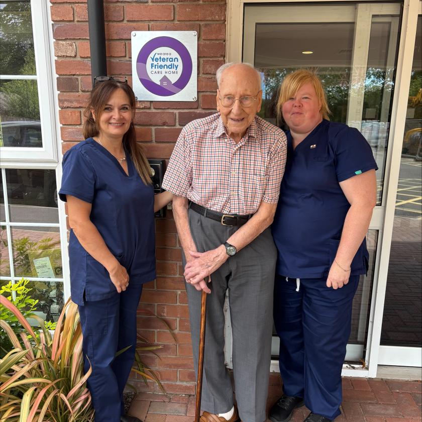 An elderly man stands in the center, holding a cane, and smiling. He is flanked by two female caregivers, both wearing navy blue scrubs.