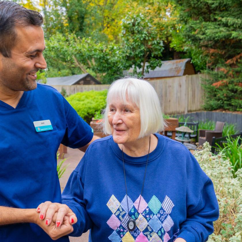A healthcare worker in a blue uniform smiles and holds hands with an elderly woman in a blue sweater adorned with a diamond pattern. They are standing in a lush garden with greenery and outdoor seating in the background. The scene conveys warmth and companionship.