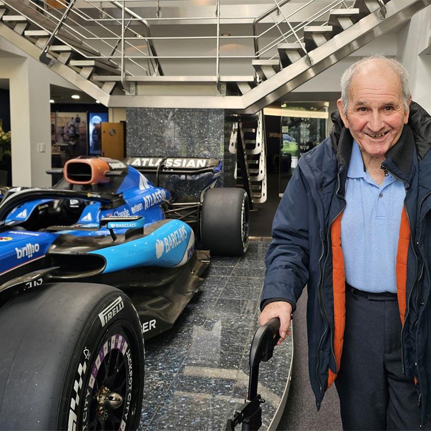 An elderly man stands next to a vibrant blue racing car, looking pleased and engaged.