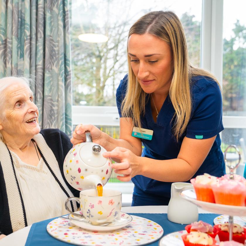A caregiver in a blue uniform is pouring tea from a decorative teapot into a cup held by an elderly woman with white hair, who is smiling and looking up at her. The setting is bright and cheerful, with a table featuring a plate of colorful desserts in the background. The atmosphere conveys warmth and companionship.