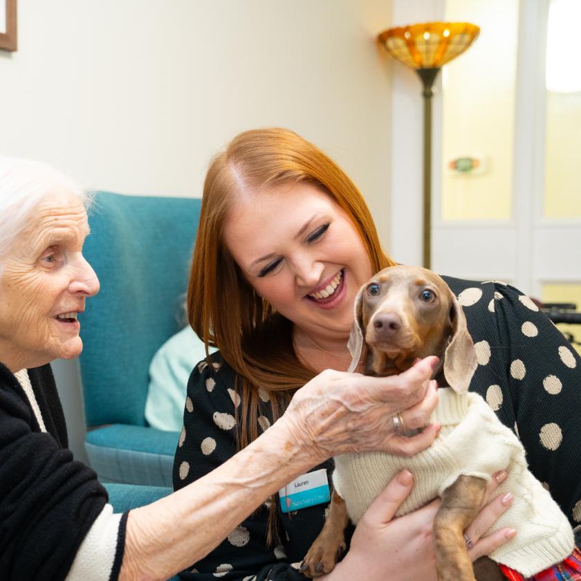 An elderly woman with white hair smiles as she interacts with a young woman who has long red hair. The young woman is holding a small dog wearing a sweater.