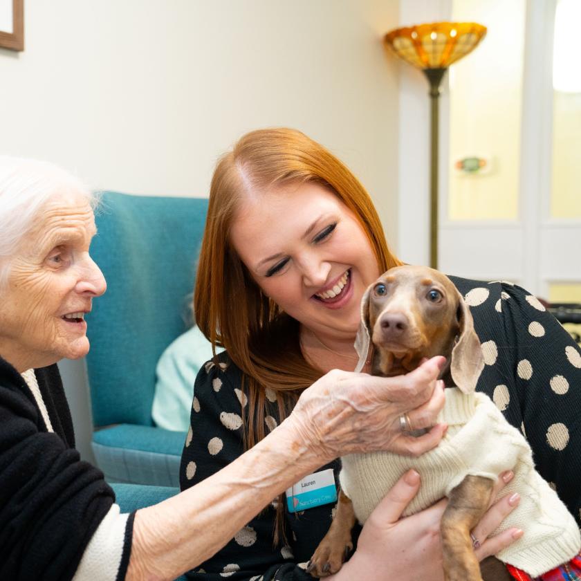 An elderly woman with white hair smiles as she interacts with a young woman who has long, reddish-brown hair. The young woman is holding a small dog wearing a sweater, and both women are laughing and enjoying the moment. They are seated in a cozy room with teal chairs and warm lighting, creating a cheerful atmosphere.