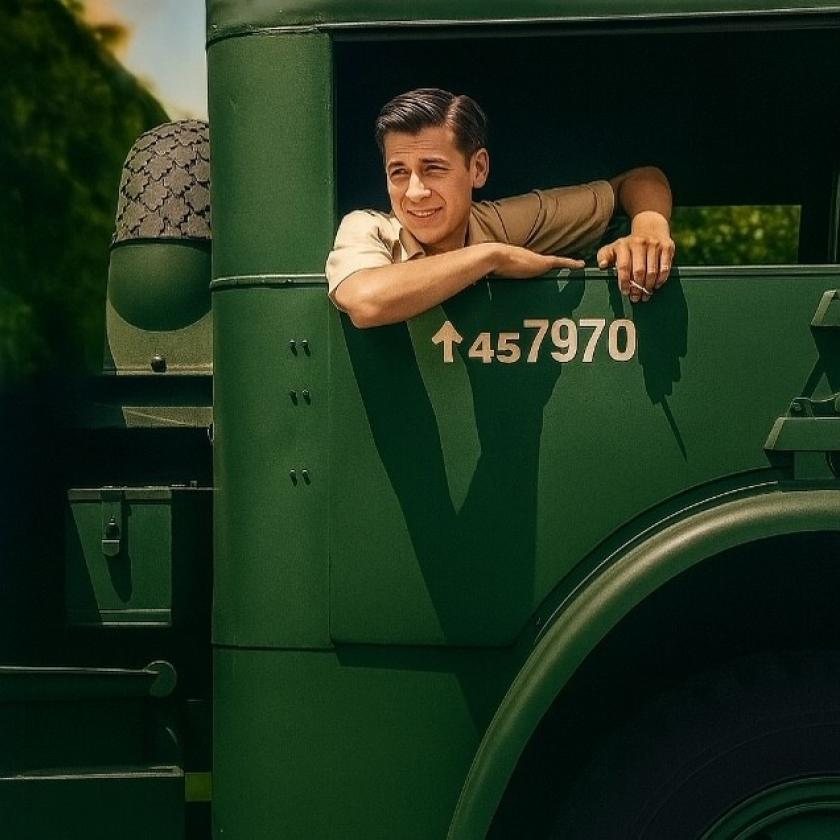 A person in a beige shirt leans out of the window of a green military vehicle, smiling. The vehicle has the number "457970" painted on its side. The background features greenery and a clear sky.