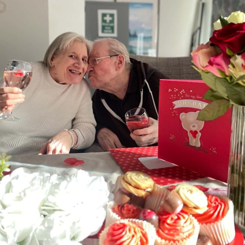 An elderly couple is sitting closely together at a table, smiling and enjoying drinks. The woman is holding a glass and leaning towards the man, who is giving her a kiss on the cheek. The table is decorated with a bouquet of flowers, cupcakes, and a large greeting card featuring a teddy bear and the word "Valentine." A red balloon is visible in the background, adding to the festive atmosphere.