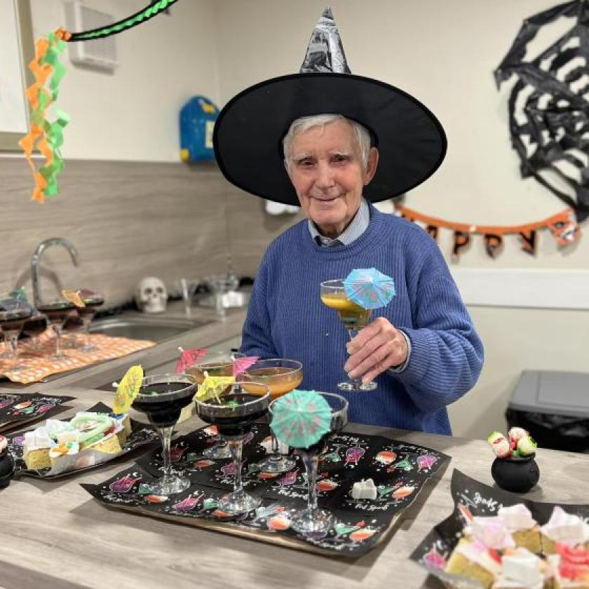 An older man in a witch hat smiles while holding a drink, embodying a festive Halloween spirit.
