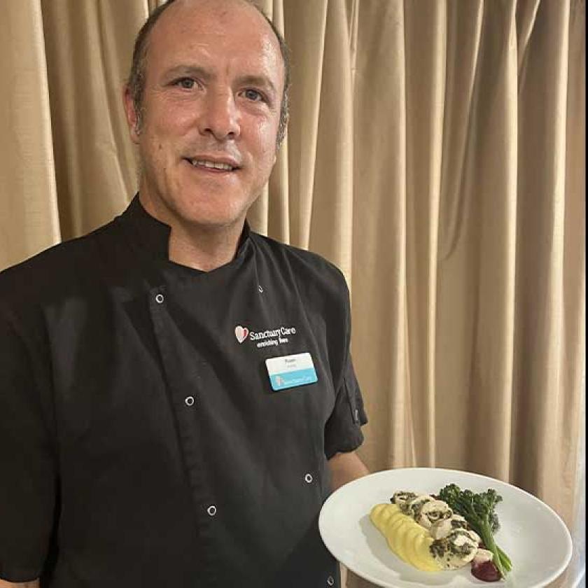 A chef wearing a black uniform with the "Sanctuary Care" logo is presenting a plate of food.