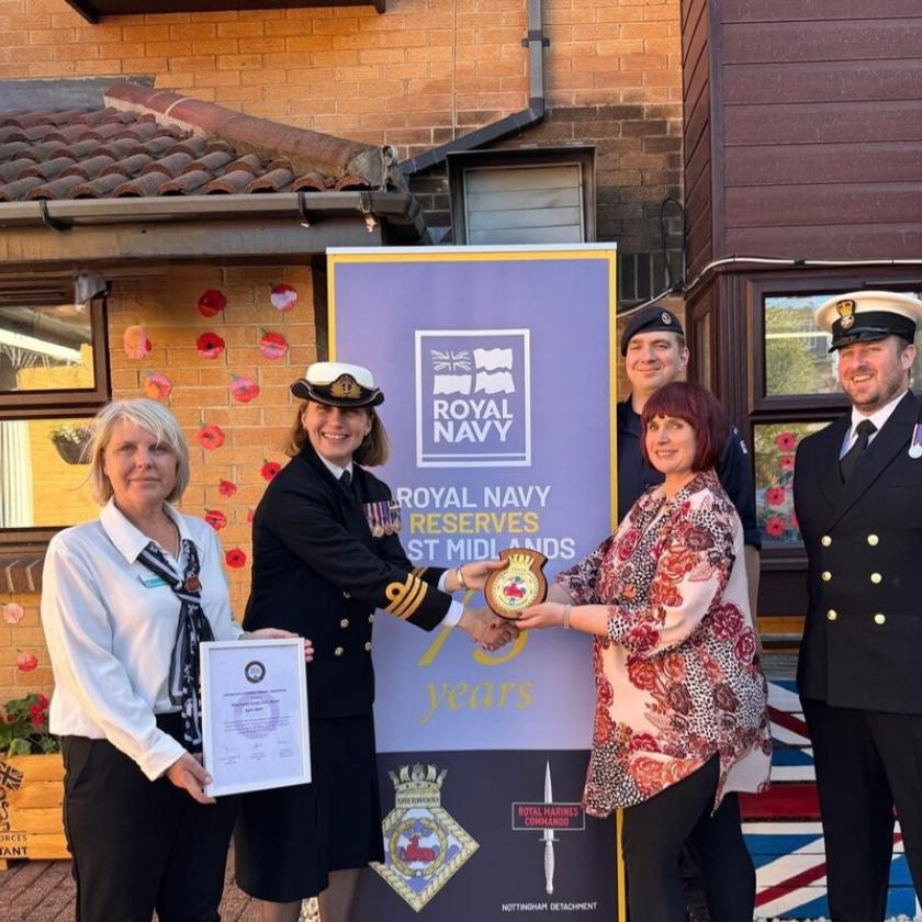 A group of five individuals stands in front of a banner celebrating 75 years of the Royal Navy Reserves in the West Midlands. The banner features the Royal Navy logo and is decorated with a Union Jack motif. Two members of the group are in naval uniforms, while the others are dressed in civilian attire. They are holding a commemorative plaque and a badge. The background includes a brick building and decorative elements, such as red paper poppies.