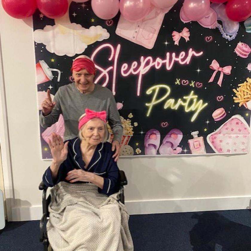 Jim and Ina Sleepover A person seated in a wheelchair and covered with a blanket poses in front of a pink-themed "Sleepover Party" backdrop, accompanied by another person standing behind. The display features pink balloons, neon-style lettering, and illustrations of sleepover items including slippers, bows, snacks, and beauty products.