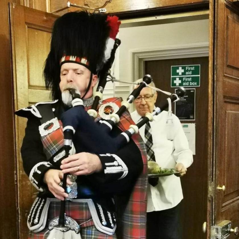 A man playing bagpipes in a kilt and a resident walking behind him